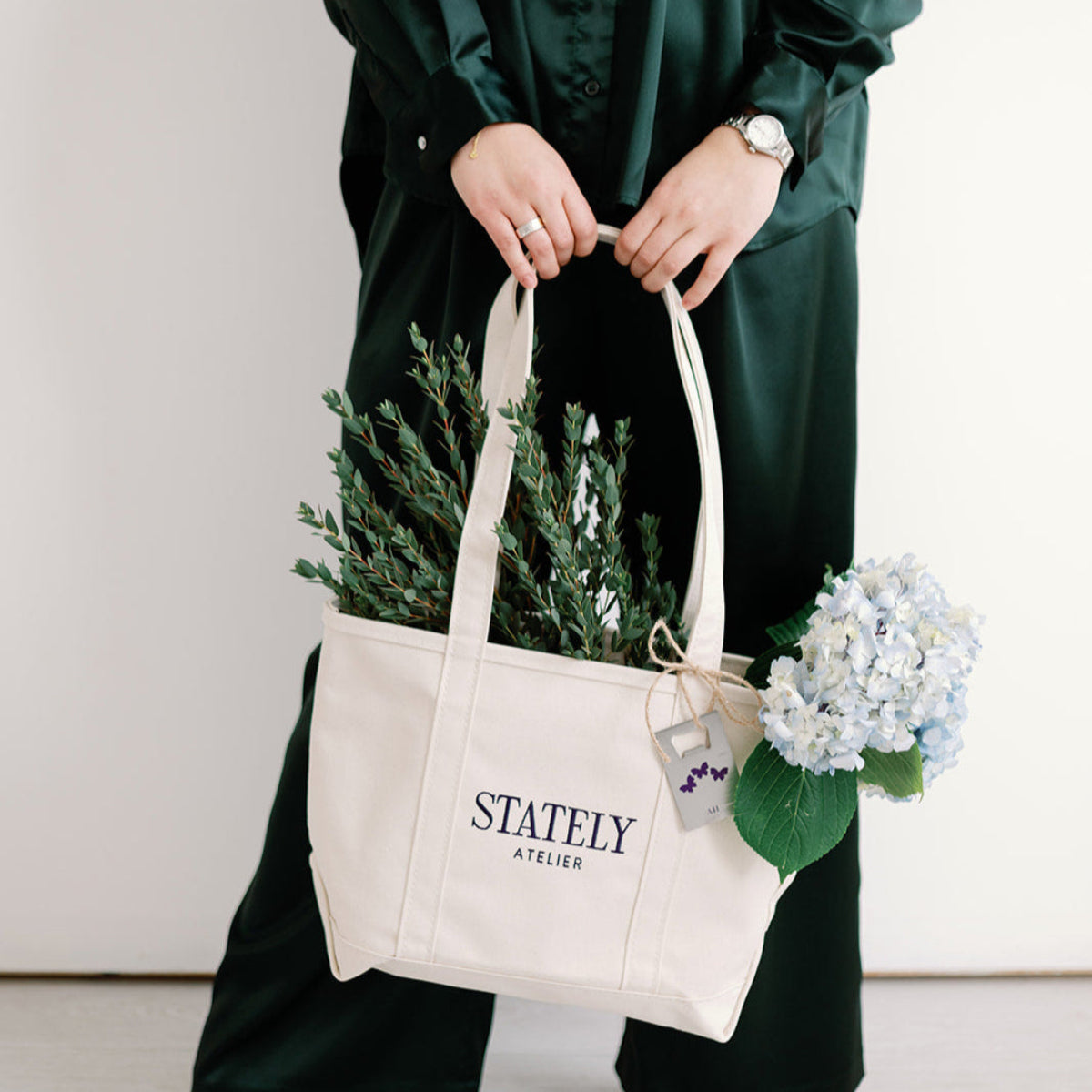 Person holding a beige tote bag with 'Stately Atelier' branding, containing plants and flowers.